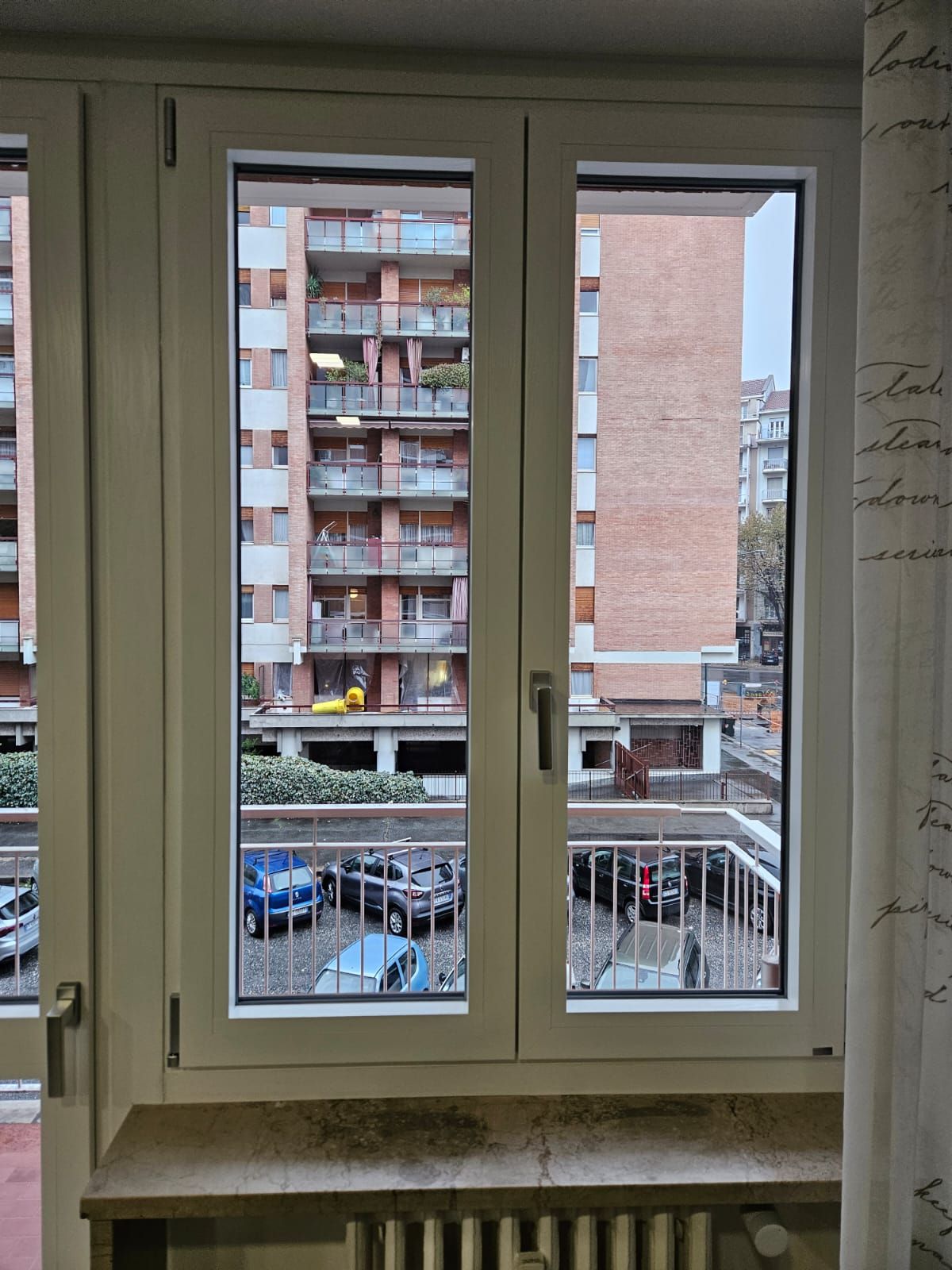 Vista dalla finestra di condomini, parcheggi e cielo coperto.