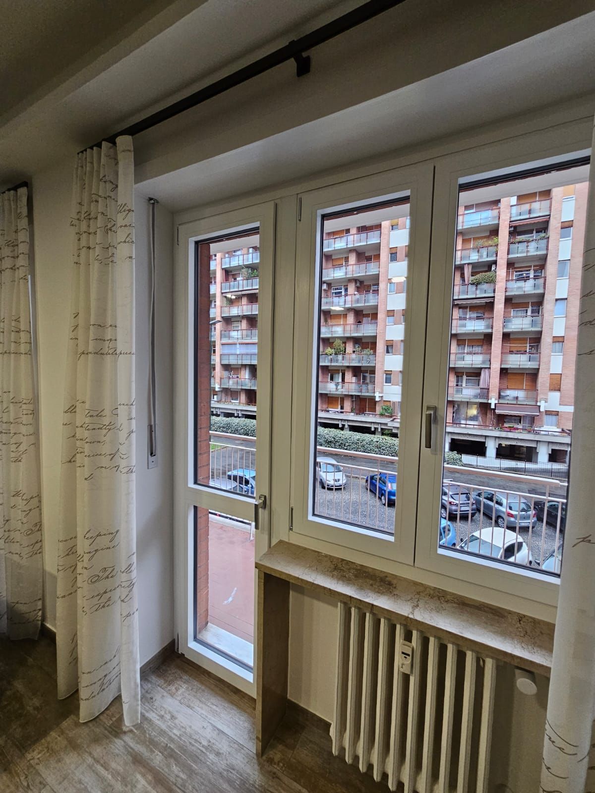 Vista dalla finestra: cornici bianche, termosifone color cuoio, tende, vista su condomini e auto parcheggiate.