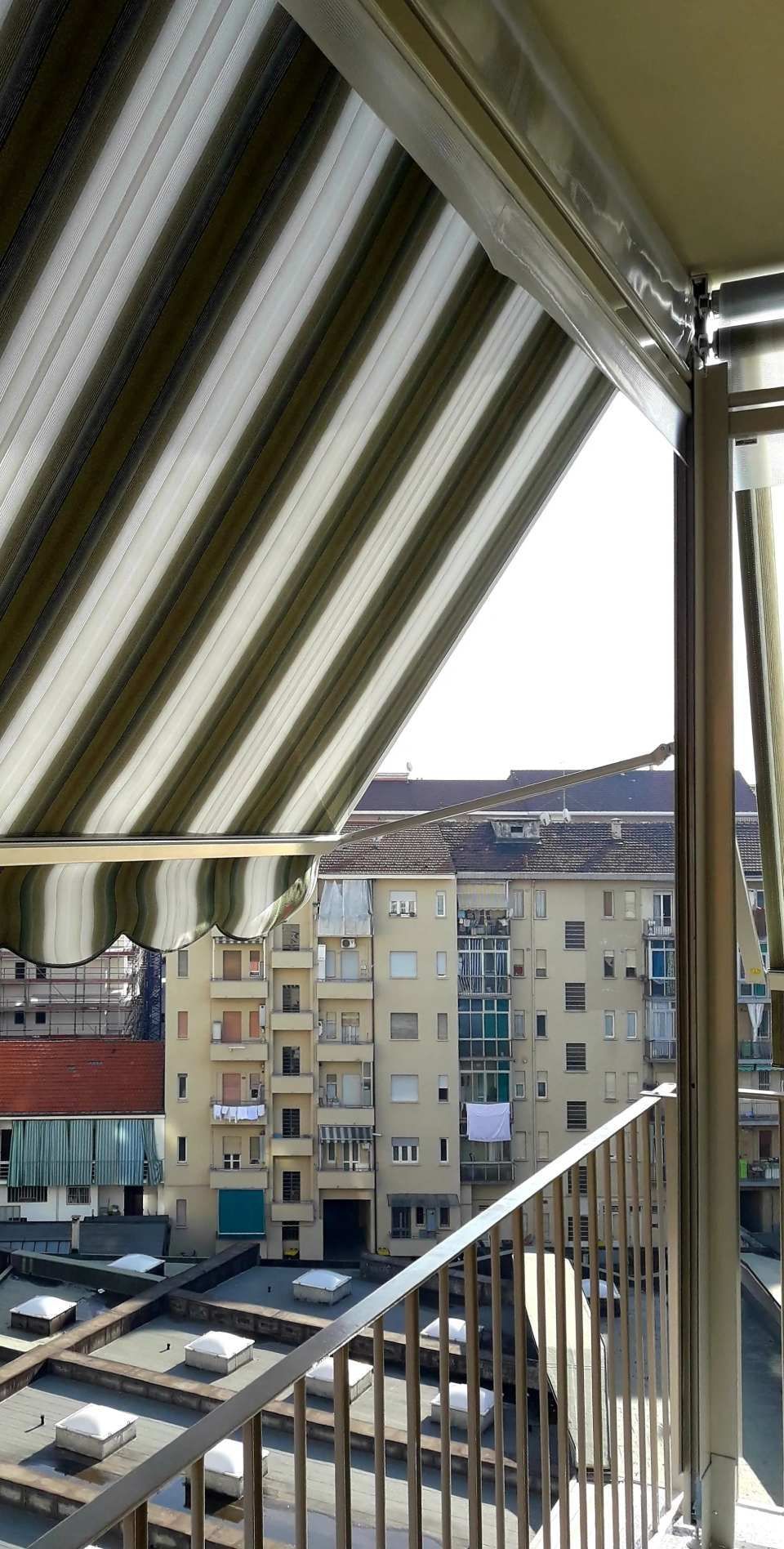 Tende da sole a strisce verdi e bianche ombreggiano un balcone che si affaccia sugli edifici cittadini.
