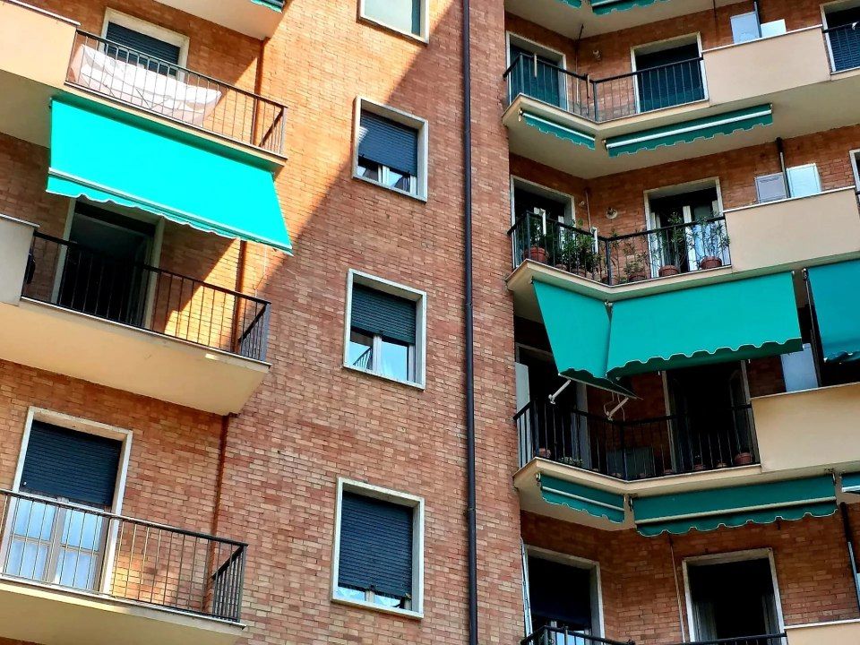 Esterno di un condominio con facciata in mattoni, balconi neri e tende da sole turchesi.