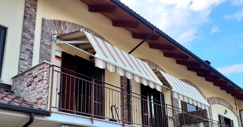 Balcone con tende da sole, ringhiera marrone, dettagli in mattoni, tende da sole a strisce beige e bianche, cielo azzurro.