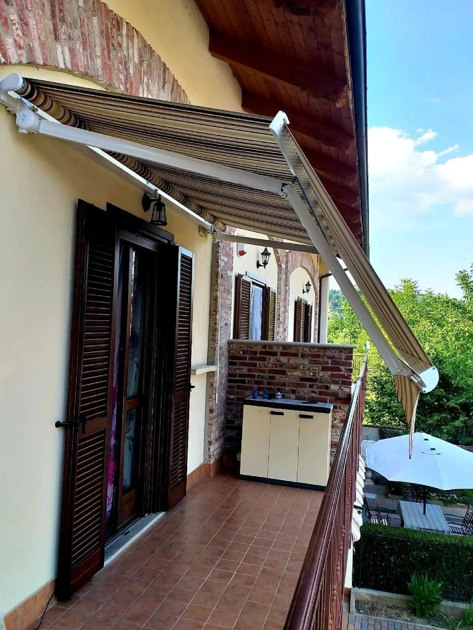 Balcone con tenda da sole beige estesa sopra la porta, muro di mattoni e vista sul giardino.