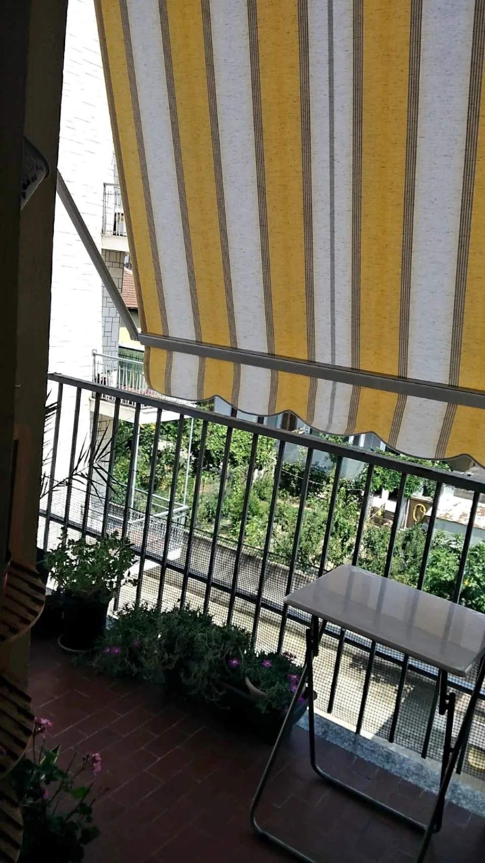 Balcone con tenda da sole a strisce gialle e bianche, ringhiera in metallo, piante in vaso e una sedia pieghevole bianca.