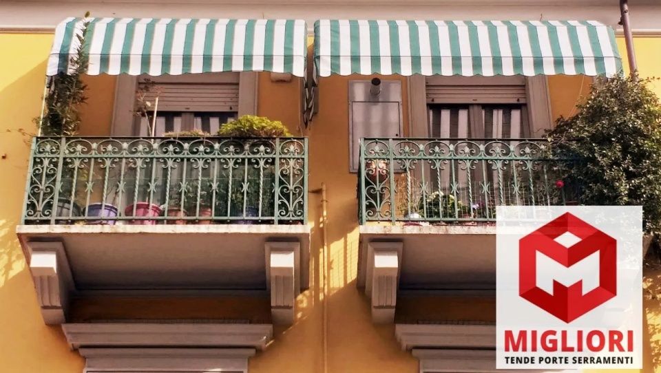 Due balconi con tende da sole a strisce verdi e bianche, ringhiere in ferro e piante su un edificio giallo.
