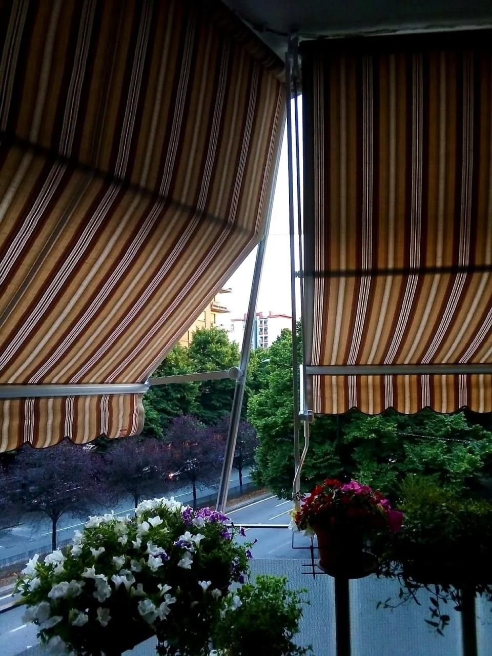 Balcone con tende a strisce, piante fiorite e vista sulla strada attraverso una vegetazione lussureggiante.