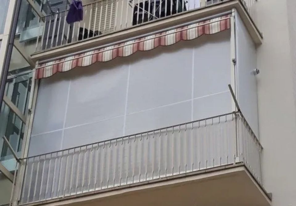 Balcone con tenda da sole a strisce bianche e rosse, pannelli laterali in plastica trasparente e ringhiera in metallo.