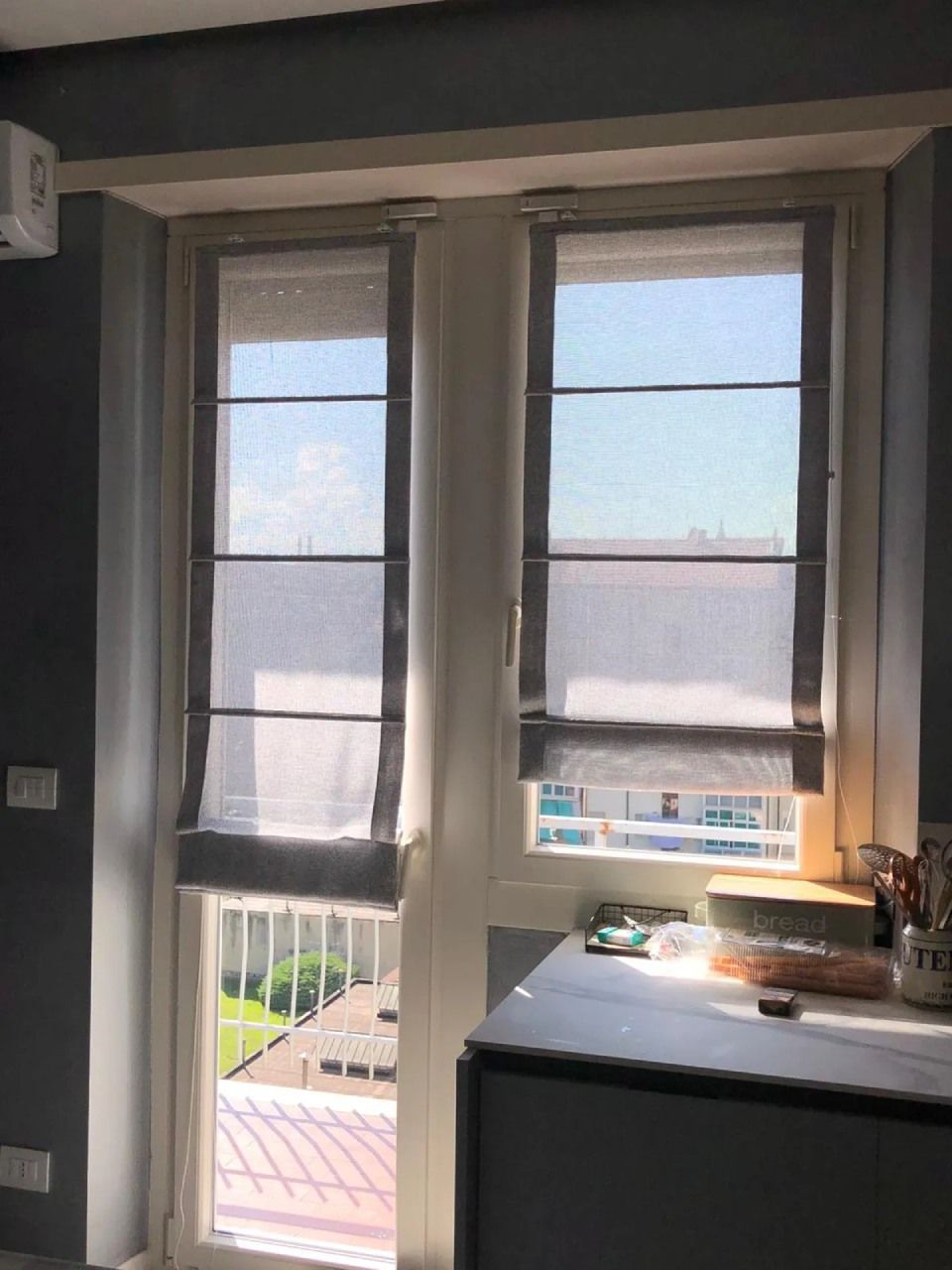 Due finestre con tende alla romana; una vista soleggiata dall'interno di una stanza.