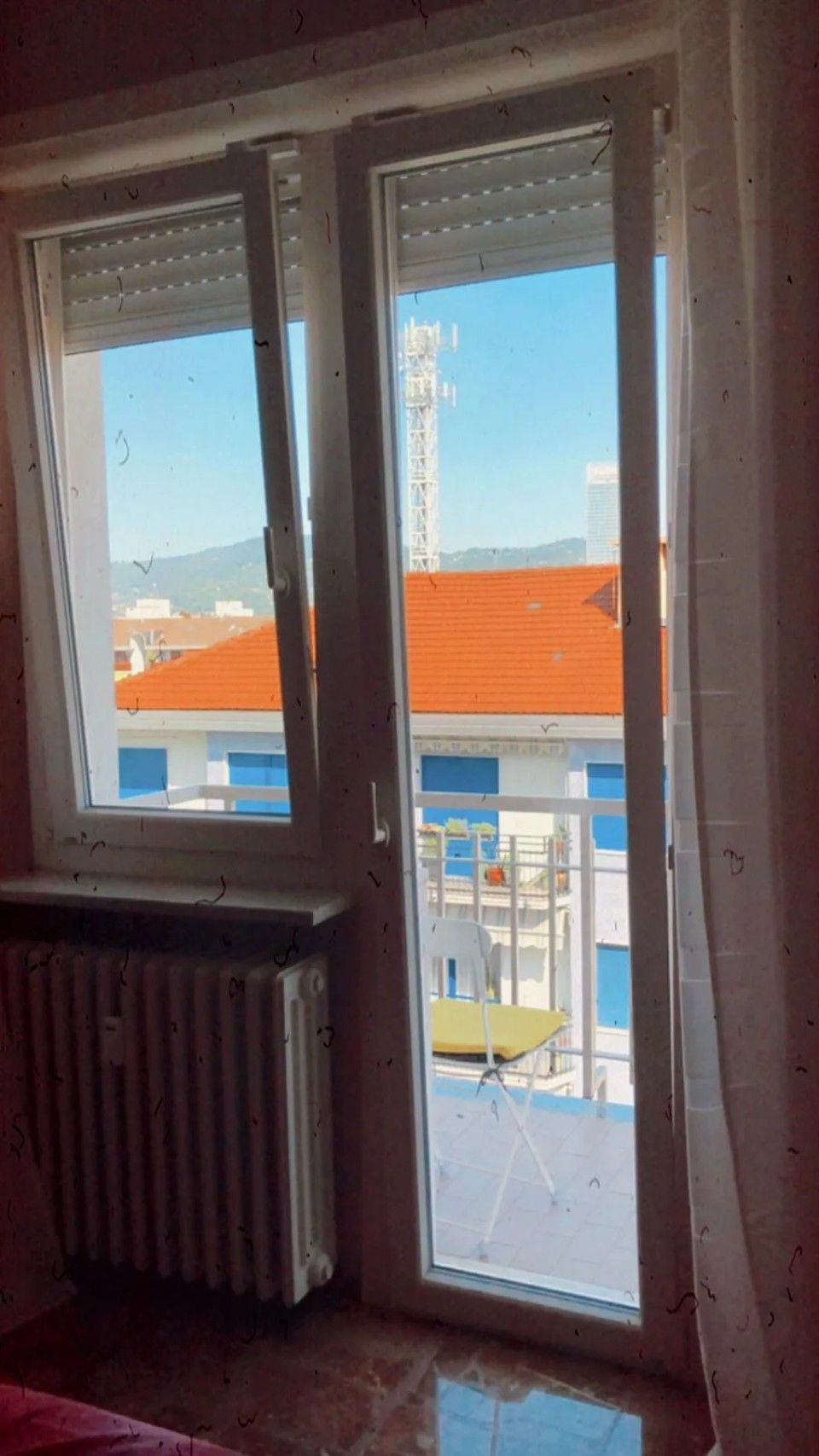 Finestra con un vetro aperto, che si affaccia su edifici con tetti rossi e un cielo azzurro brillante.