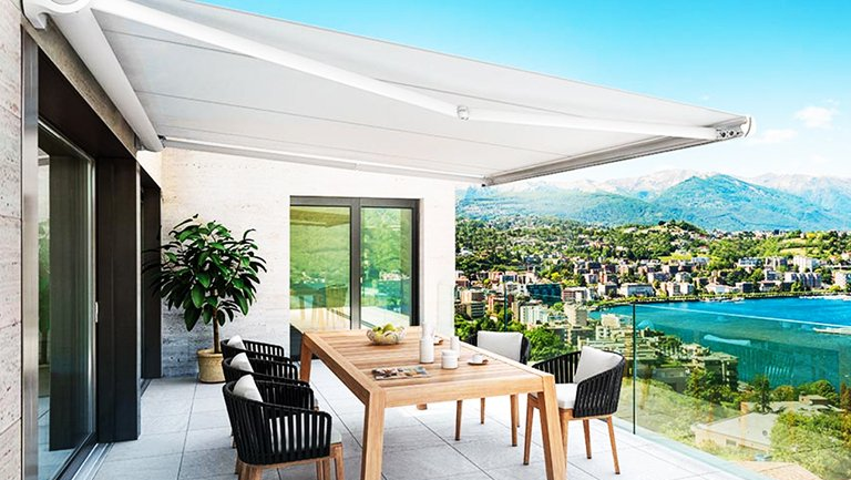 Balcone con tenda da sole bianca, tavolo da pranzo, sedie e vista sul lago e sulle montagne.