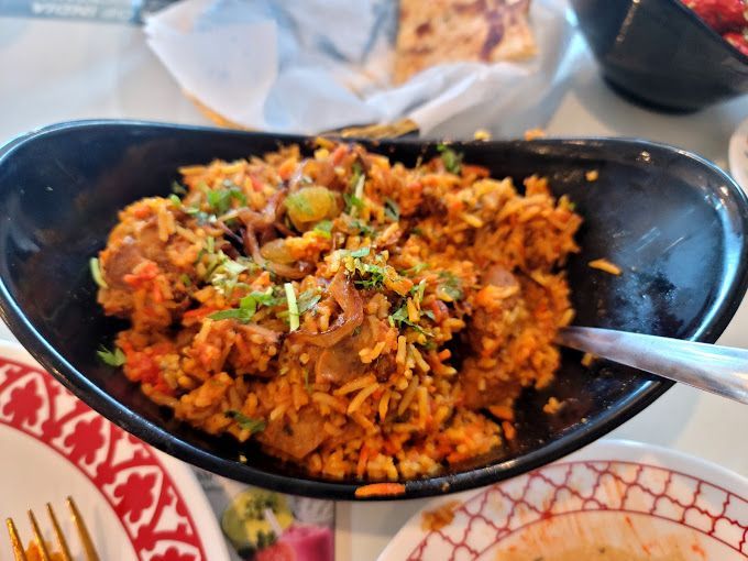Bowl of reddish-brown biryani rice, garnished with green herbs, on a table.