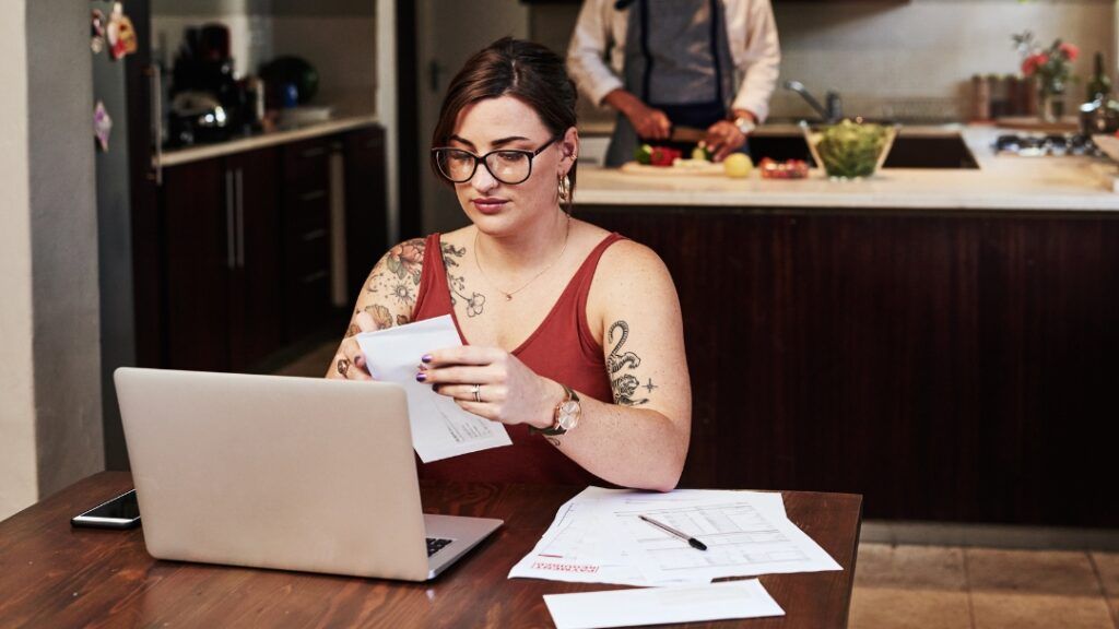 Woman at table with laptop reviews bills, man in kitchen prepares food.