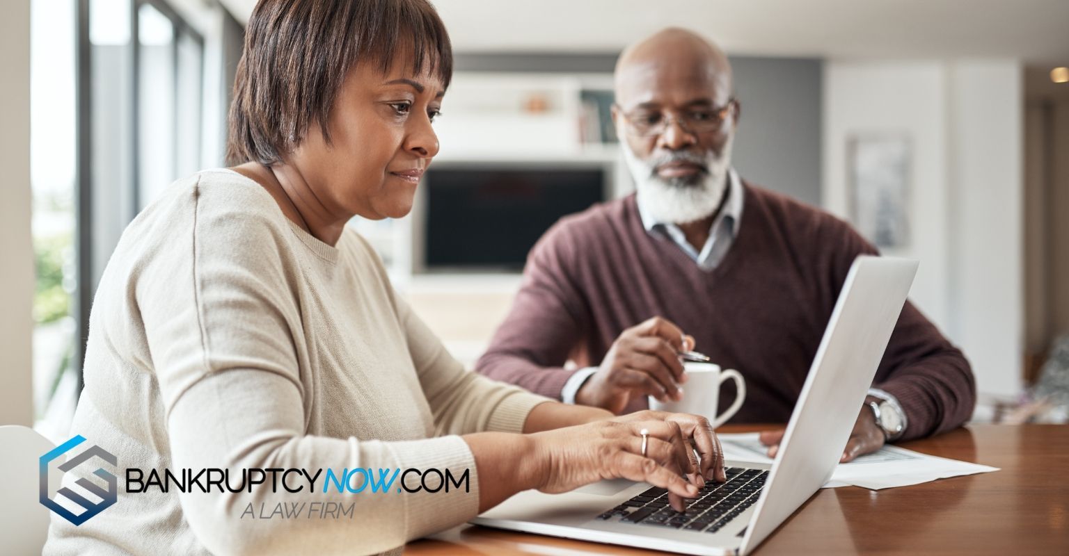 Woman using laptop, man looks on, both with serious expressions, sitting at a table. 