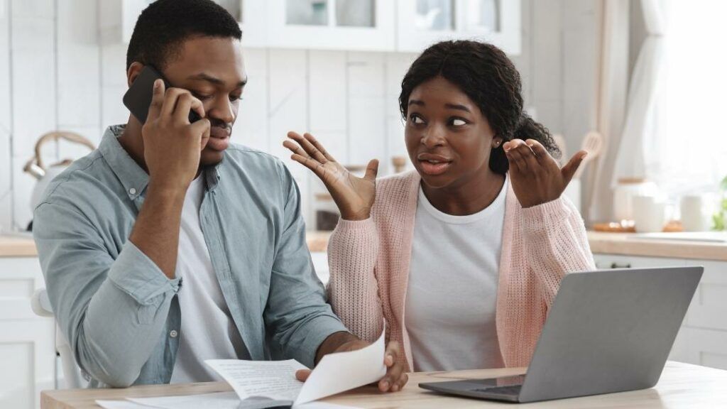 Couple reviewing bills, man on phone, woman gesturing, laptop on table.