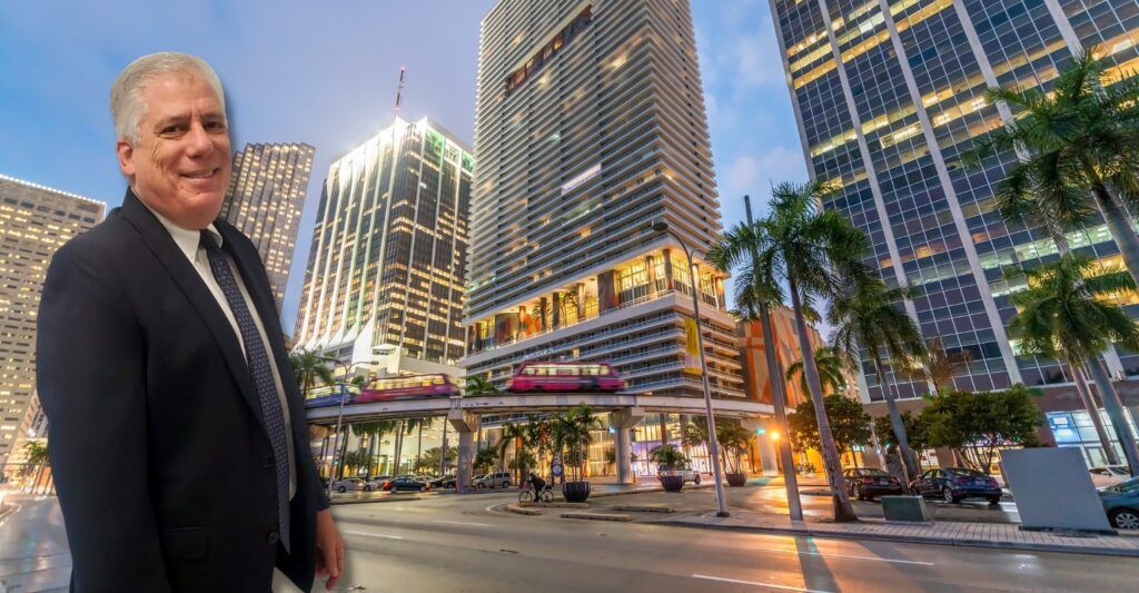 Man in suit stands in front of Miami skyline, near a raised train.