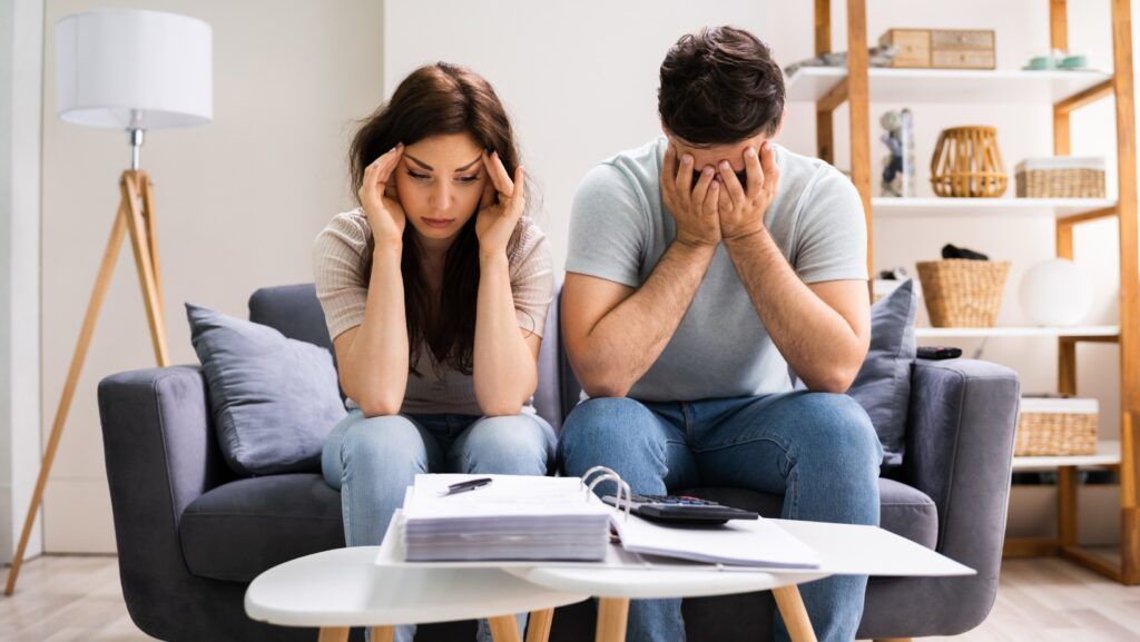 Couple on a couch stressed about finances, covering their faces. Papers and calculator on table.