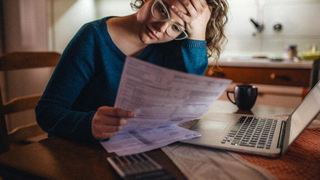 Woman with glasses looks stressed, holding bills, sitting at a table with a laptop and calculator.