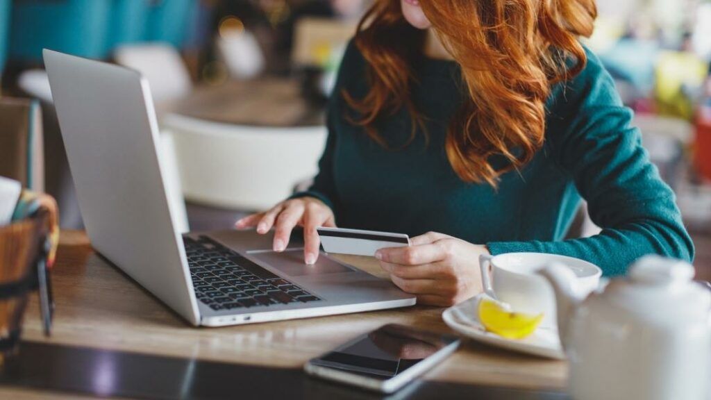 Woman with red hair using a laptop, holding a credit card, at a table with tea and a smartphone.