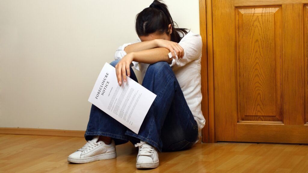 Woman seated on floor, head in arms, holding a test paper, near a door.