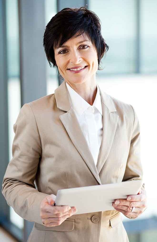 Woman in beige suit, holding a tablet, smiling, indoors, near a window.
