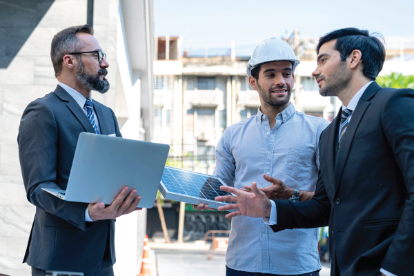 Three men discussing solar panel installation outdoors; one holds a laptop, another a panel, and the third wears a hard hat.