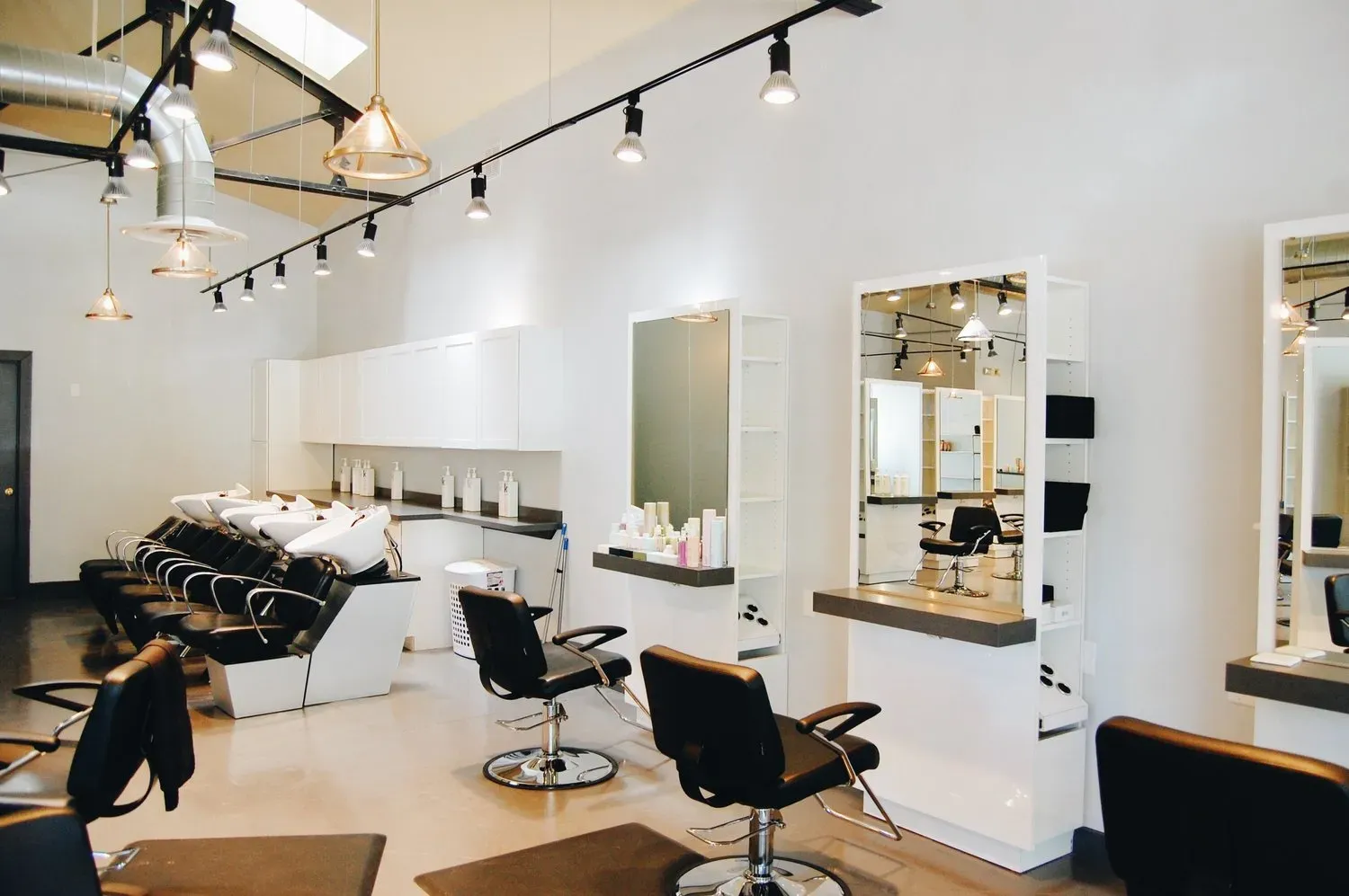 A modern, brightly lit hair salon with black styling chairs, washing stations, and white wall-mounted mirrors.