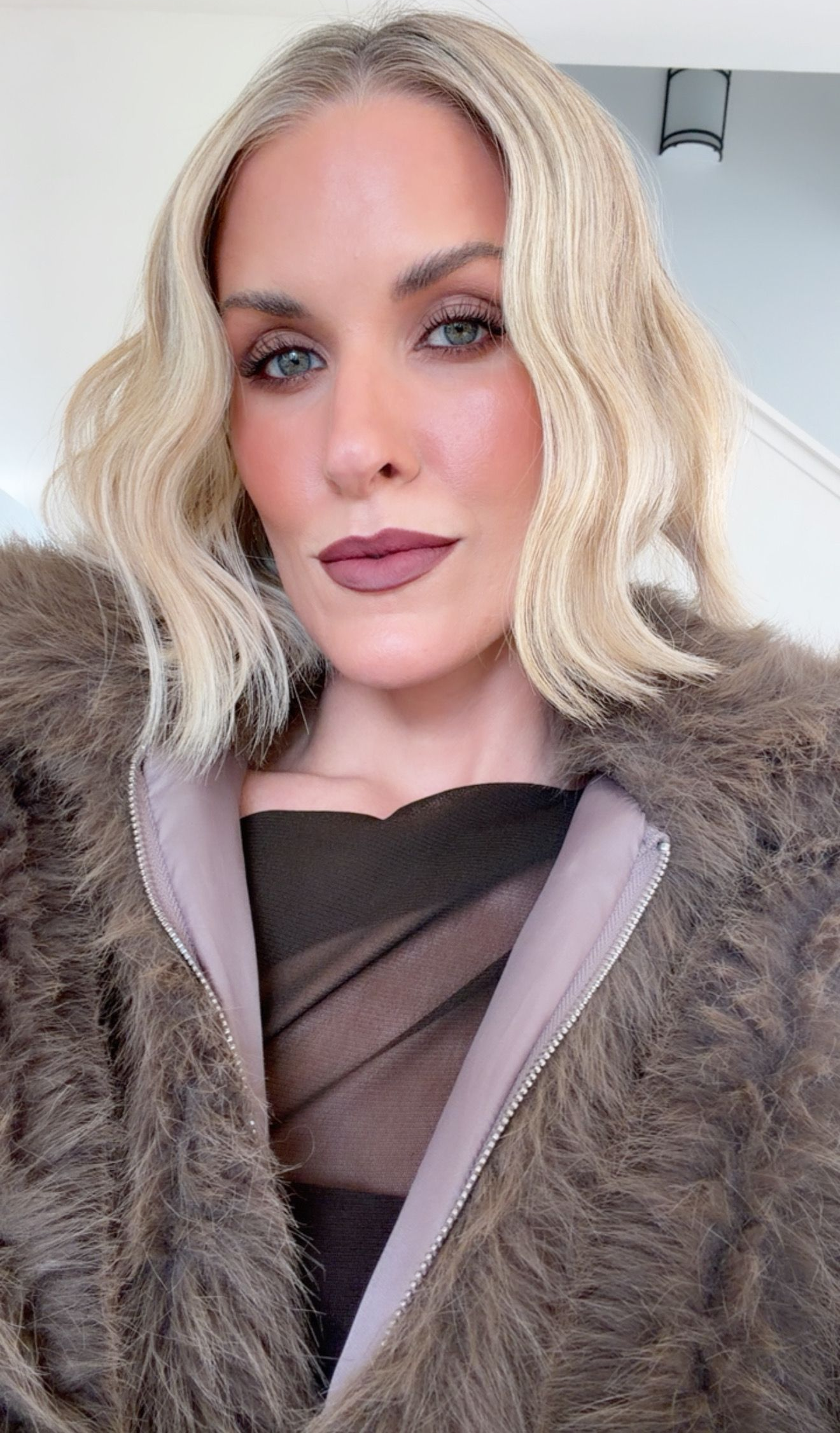A person with shoulder-length, wavy blonde hair wearing a brown faux fur coat and a dark top, looking at the camera.