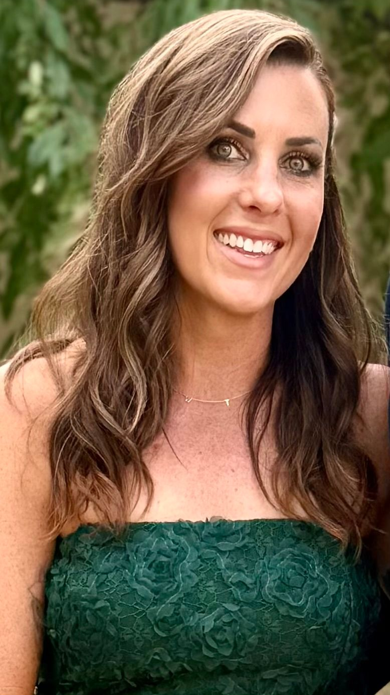 A smiling person with shoulder-length hair and a layered outfit posing against a plain background.