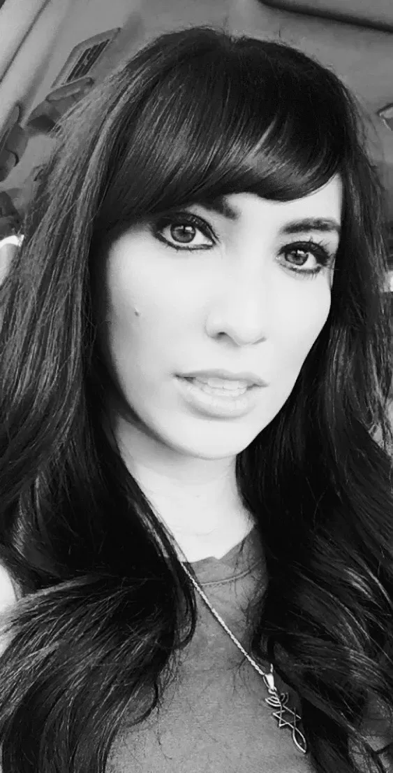 A black-and-white portrait of a person with long, dark hair and bangs, looking directly at the camera with a neutral gaze.