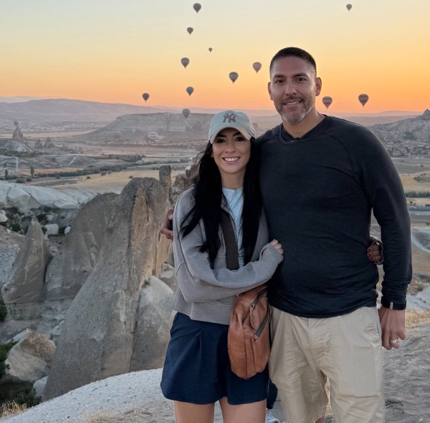 A couple stands together smiling in Cappadocia, Turkey, against a sunset landscape filled with floating hot air balloons.