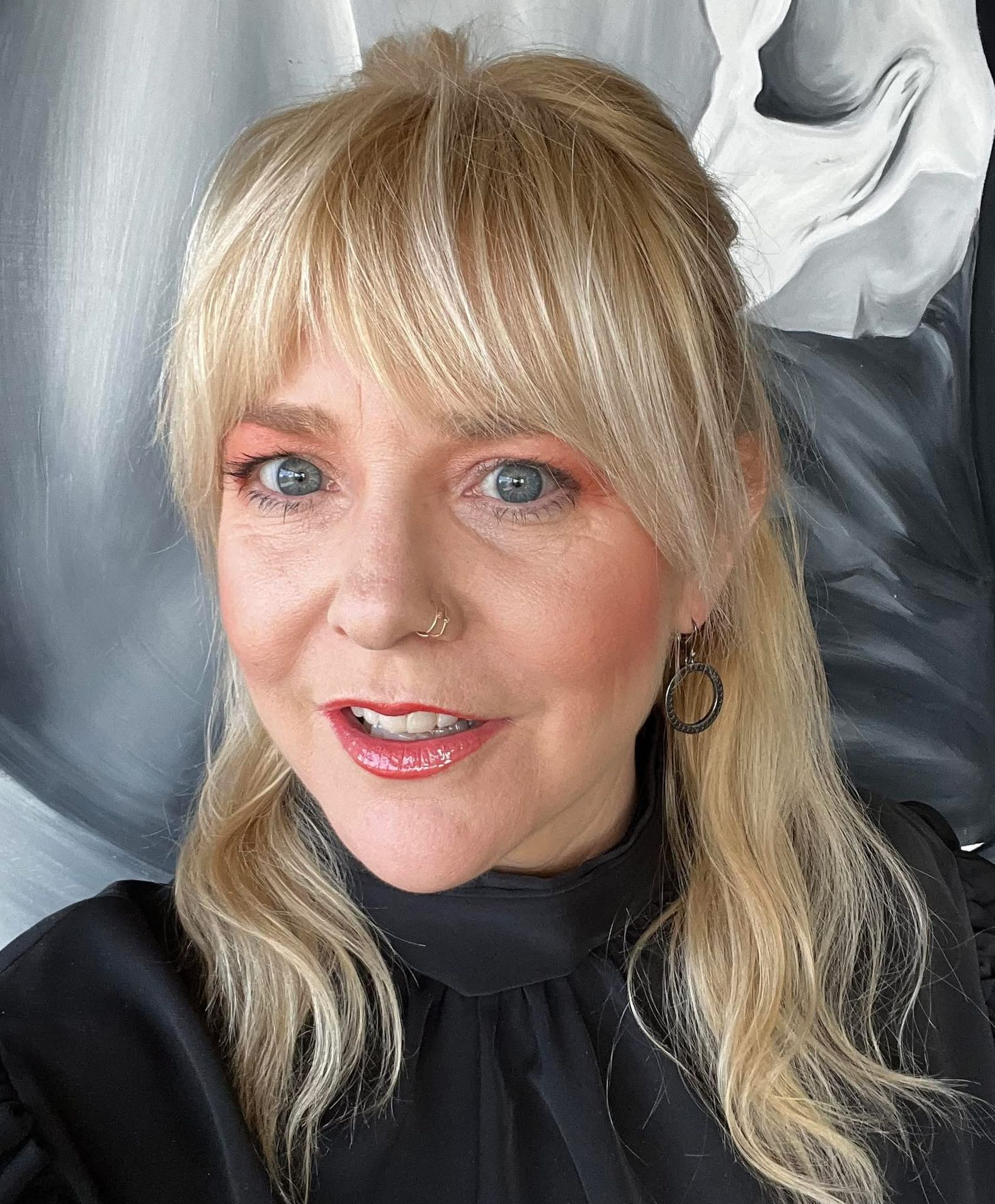 A blonde person with bangs and a nose ring wearing a black top, smiling at the camera against a neutral background.