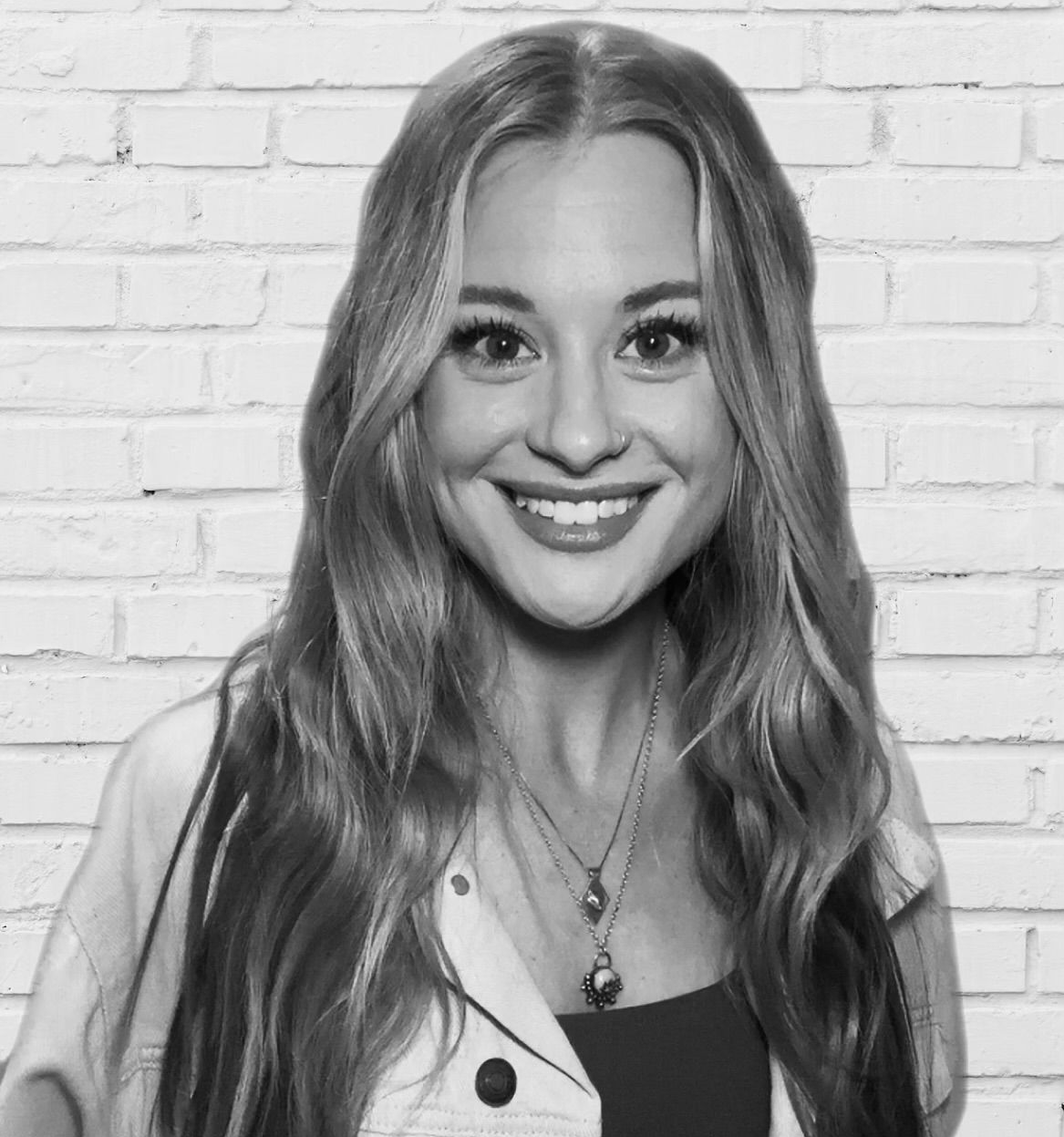 A professional black-and-white headshot of a smiling person with long wavy hair, wearing a blazer against a dark background.