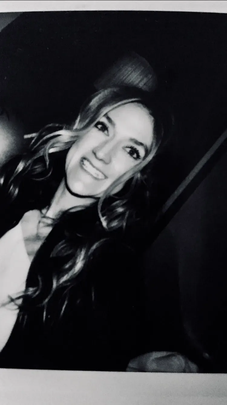 A smiling person with long hair poses for a black-and-white selfie in a dimly lit setting.
