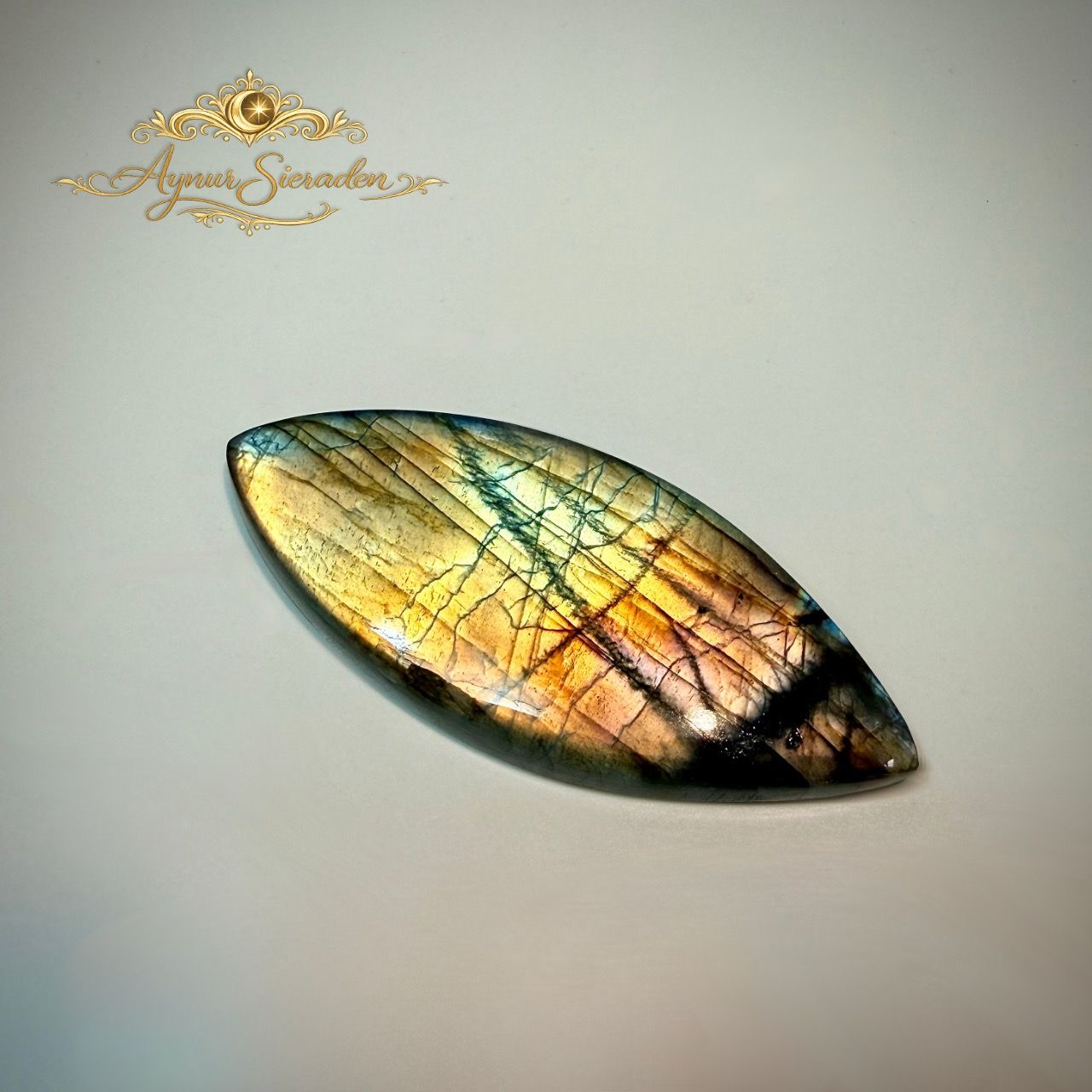 Marquisevormige labradoriet cabochon met goudgroene glans op lichte achtergrond.