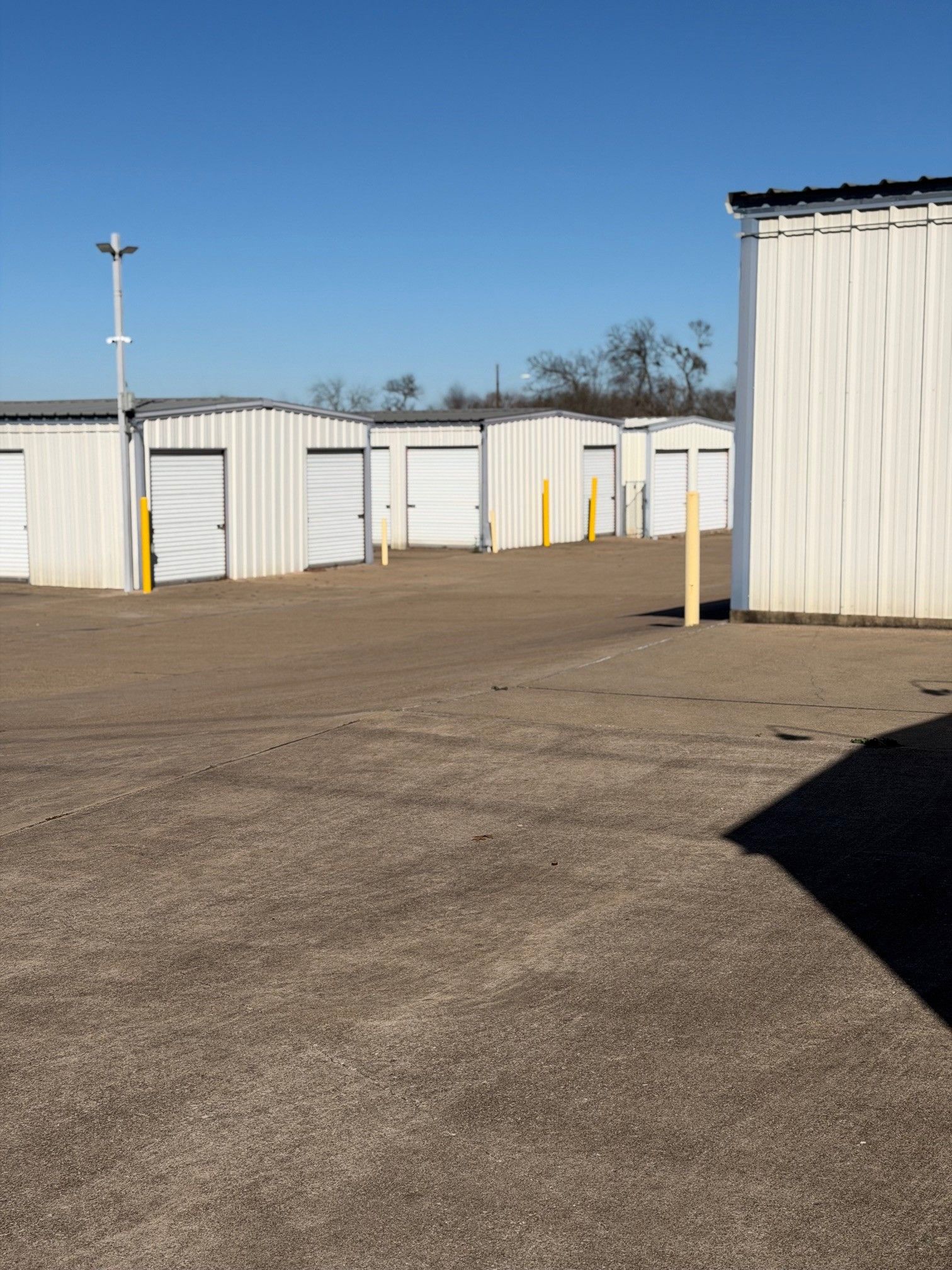 Storage units in a gravel lot under a clear blue sky. White doors, metal siding, yellow bollards.
