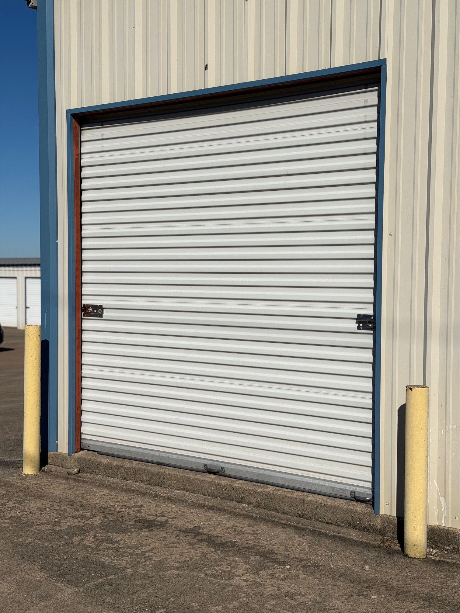 Metal roll-up storage door, closed, in a building. Two yellow posts flank it. Blue trim.