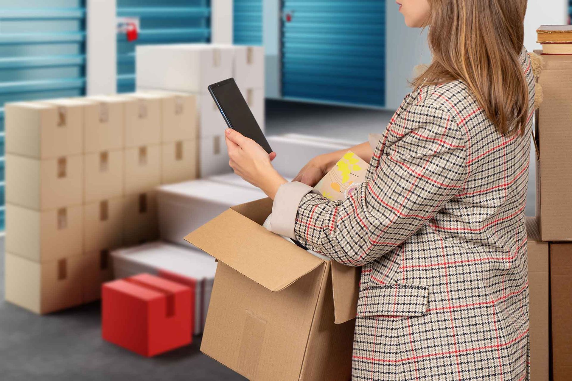 Woman in storage unit packing a box, reading a notepad. Boxes stacked behind her.