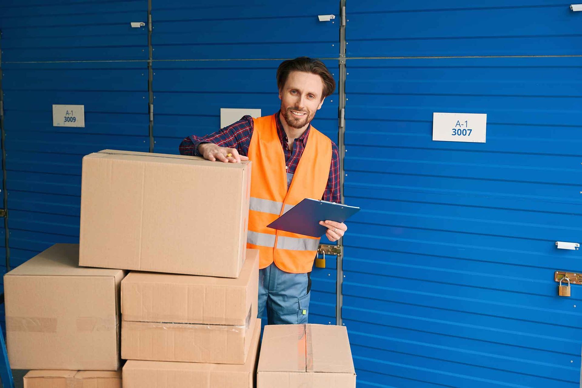 Man in orange vest and clipboard leaning on stack of boxes in front of blue storage lockers.