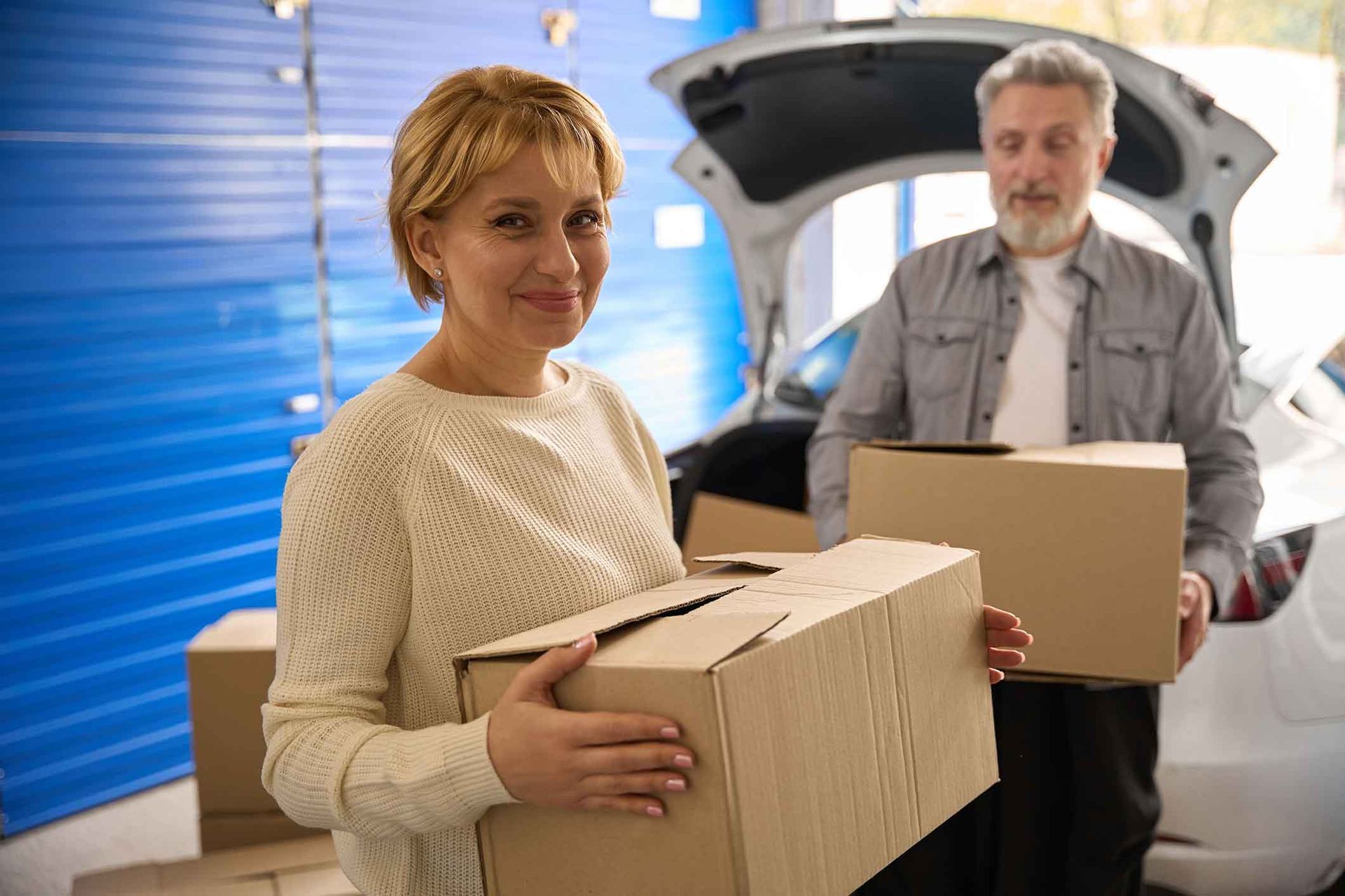 Woman and man carrying cardboard boxes near a car with the trunk open, indoors.