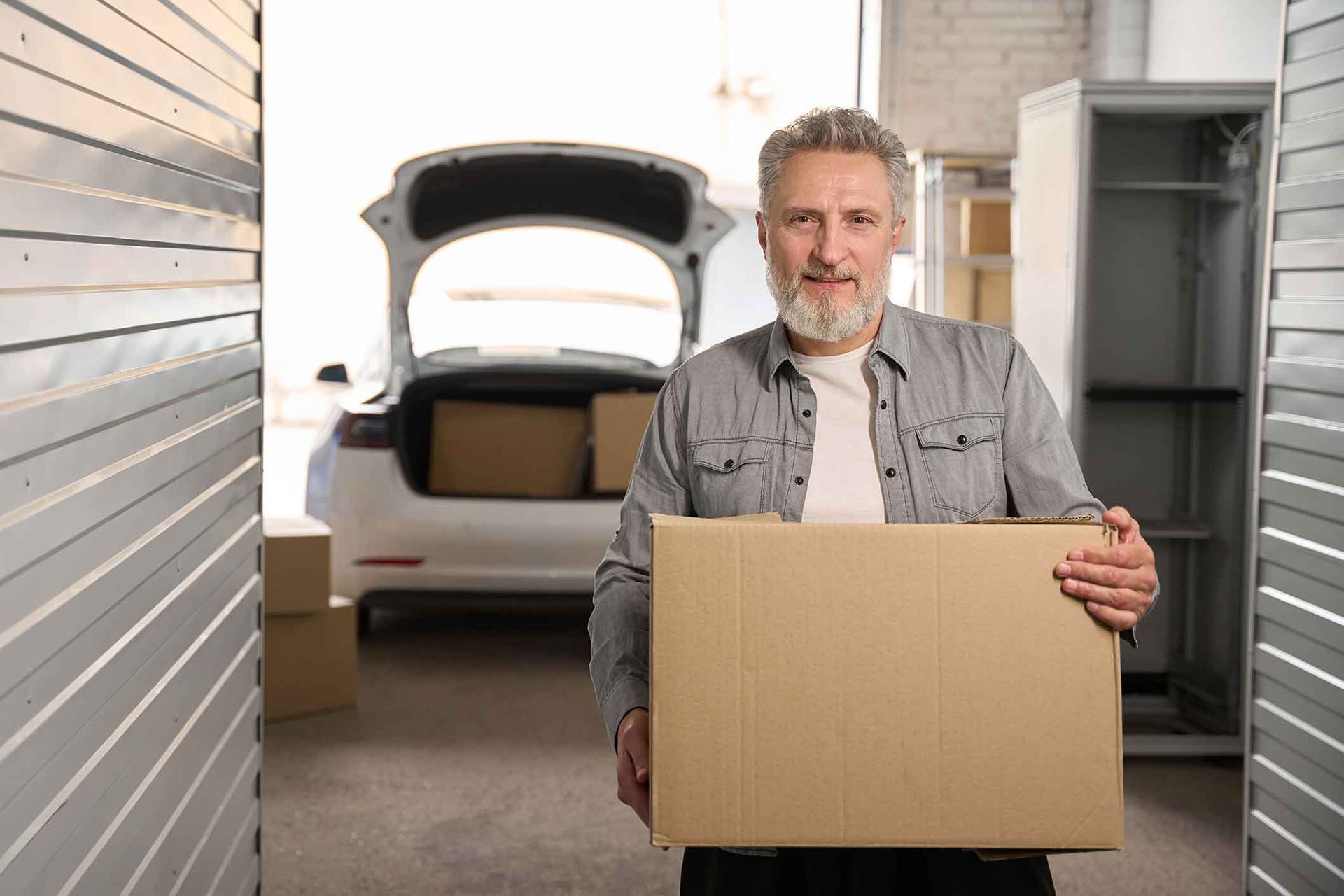 Man holding a cardboard box in front of a car with an open trunk in a storage unit.