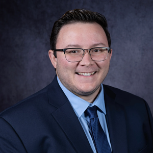 Smiling person in a navy suit and glasses against a dark studio background