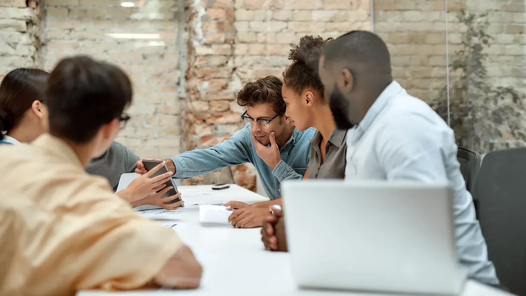 People in a meeting around a table, discussing work with laptops in a bright office.