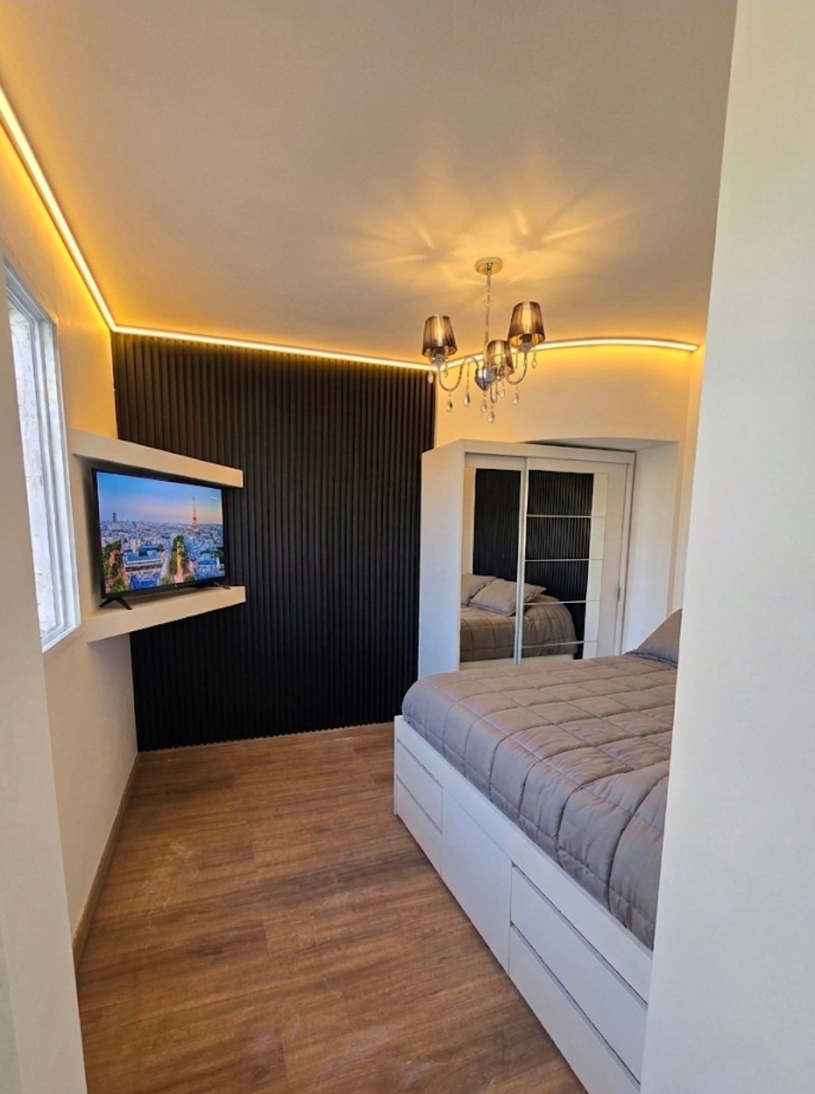 Dormitorio moderno con cama, televisor montado en la pared, pared decorativa oscura, luces cálidas en el techo