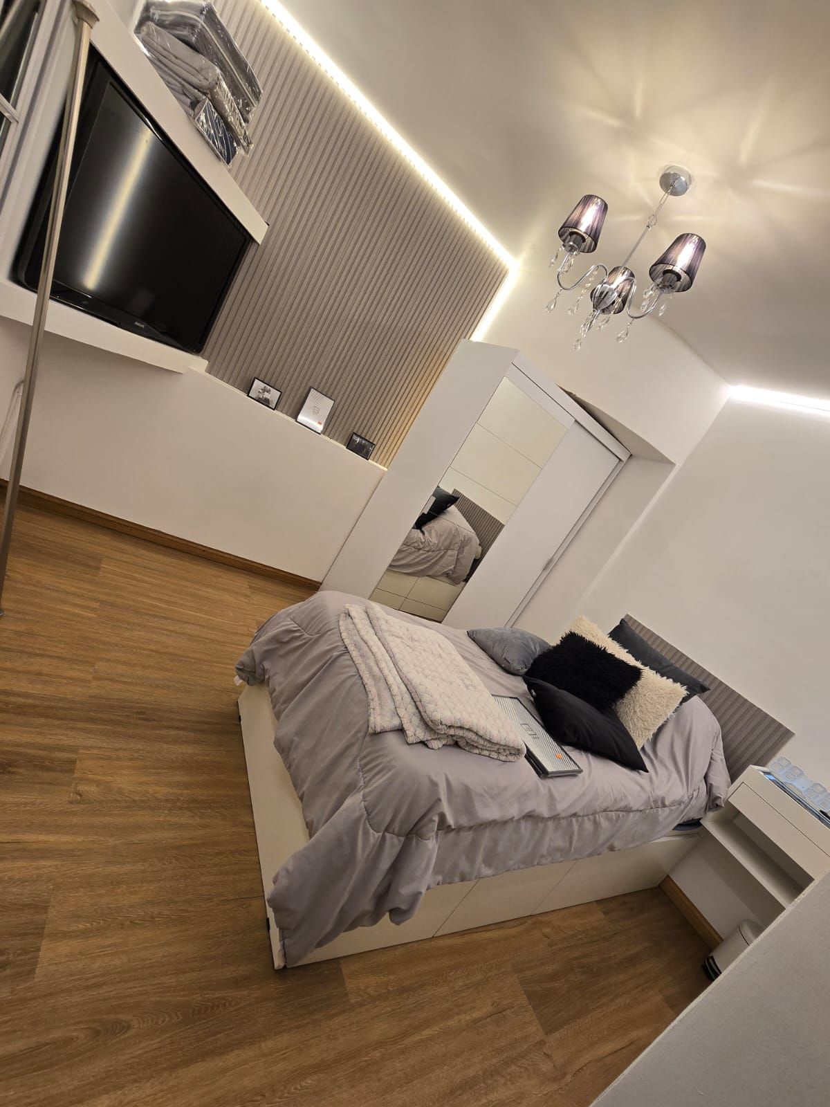 Dormitorio moderno con cama, suelo de madera, armario con espejo, televisor y lámpara de araña.