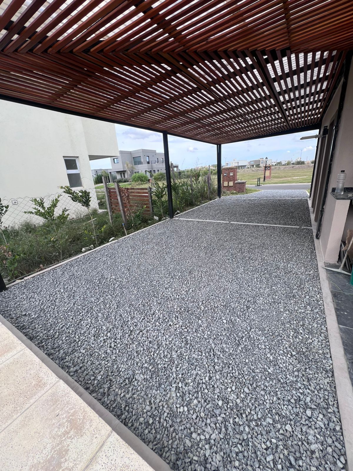 Patio cubierto de grava con techo de listones de madera junto a un edificio blanco y vistas al jardín.