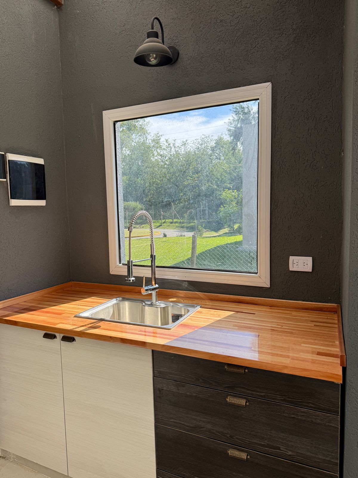 Fregadero de cocina moderno bajo una ventana con encimera de madera, paredes oscuras y vegetación en el exterior.