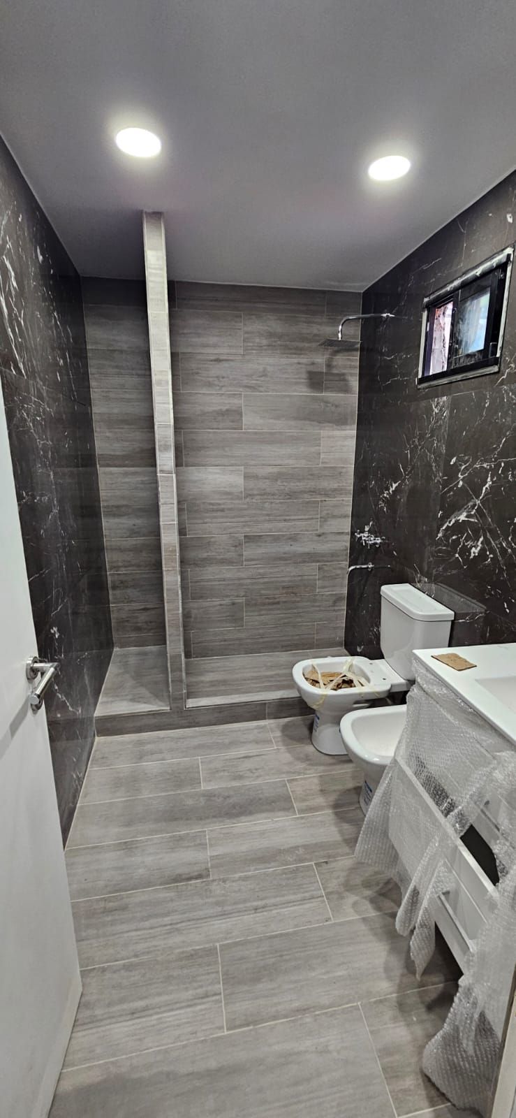 Baño moderno con azulejos grises, inodoro, lavabo, zona de ducha y luces empotradas en el techo.