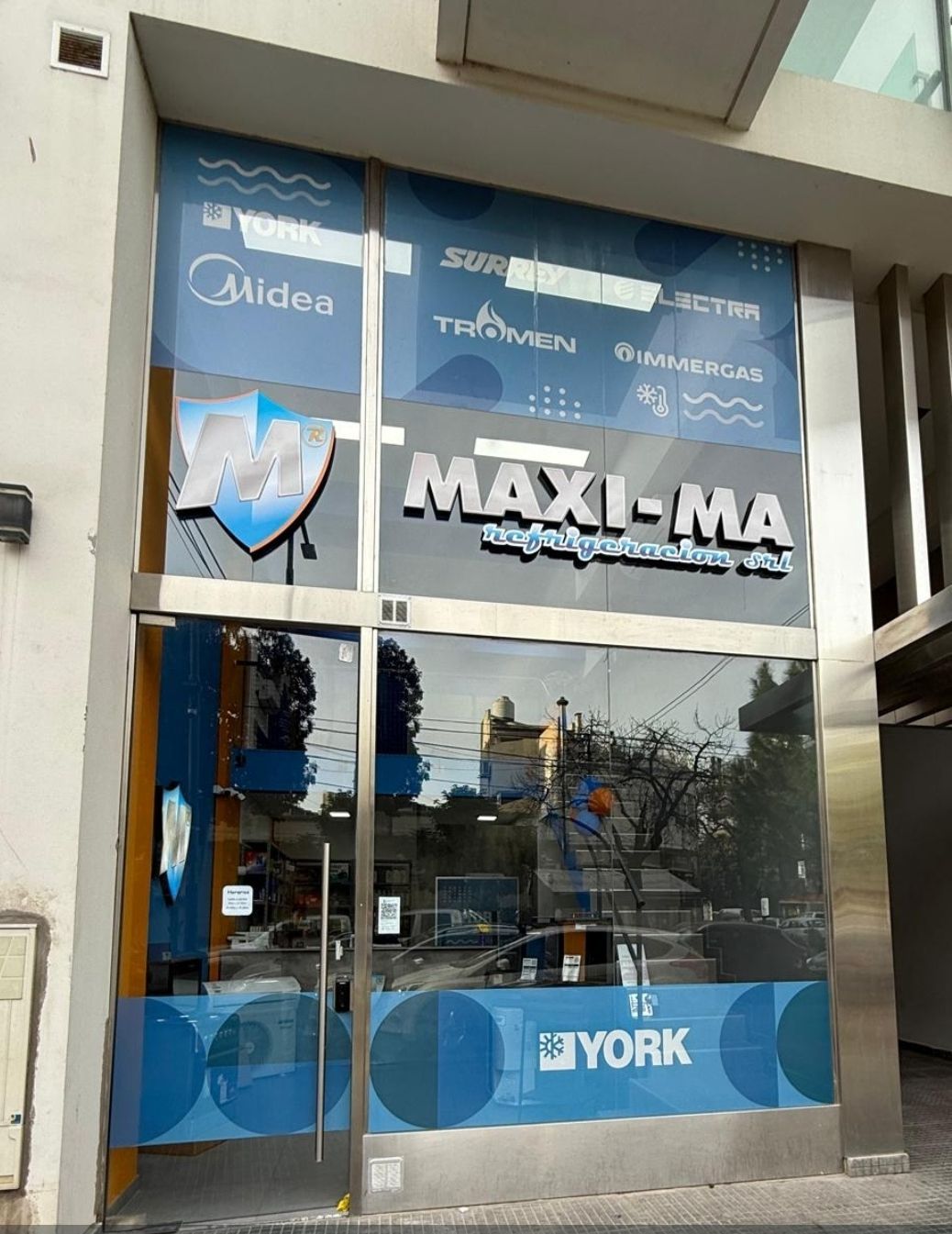 Fachada con letrero “MAXI-MA”, logotipos en color azul y logotipos de York HVAC en la entrada de cristal.