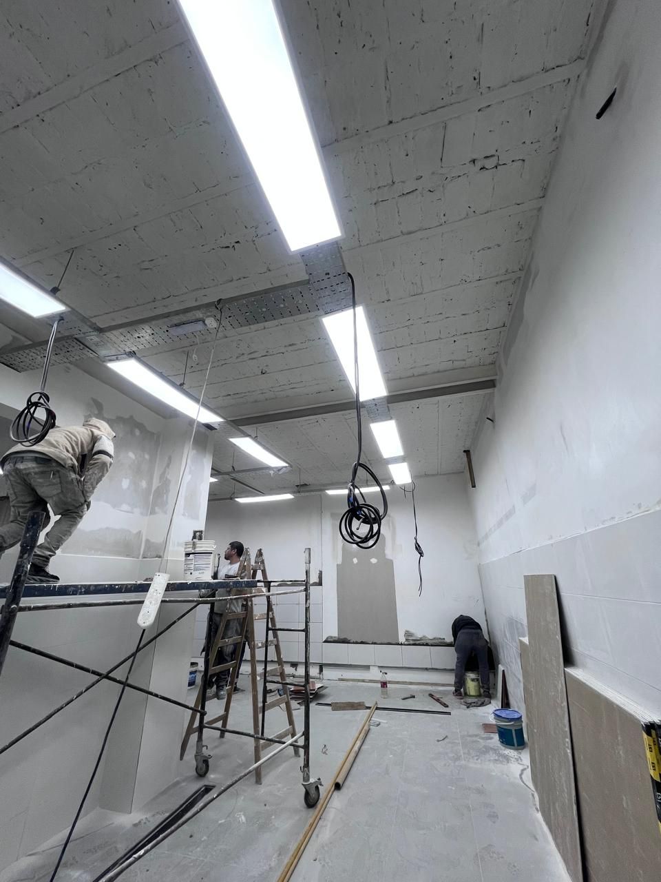 Habitación parcialmente renovada con trabajadores, andamios, luces colgantes y materiales de construcción en el suelo.