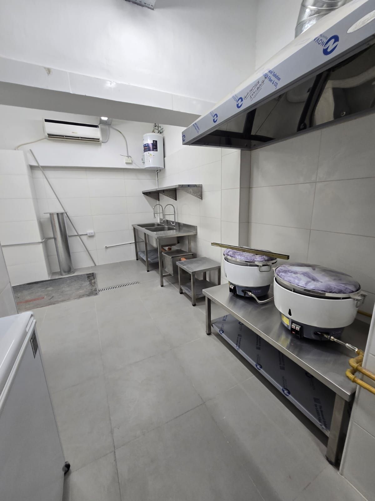 Cocina comercial vacía con encimeras, fregaderos y ollas de acero inoxidable bajo una campana extractora.