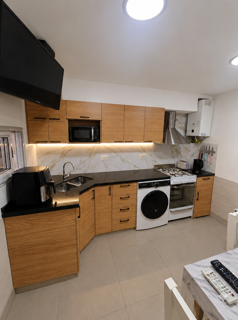 Cocina moderna con armarios de madera, encimeras negras, horno blanco y lavadora bajo luces de techo brillantes.